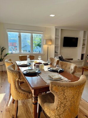 Comedor con una mesa de madera dispuesta para seis personas, rodeada de sillas tapizadas. Un televisor de pantalla plana está montado en la pared, y un ventanal ofrece vistas al exterior.