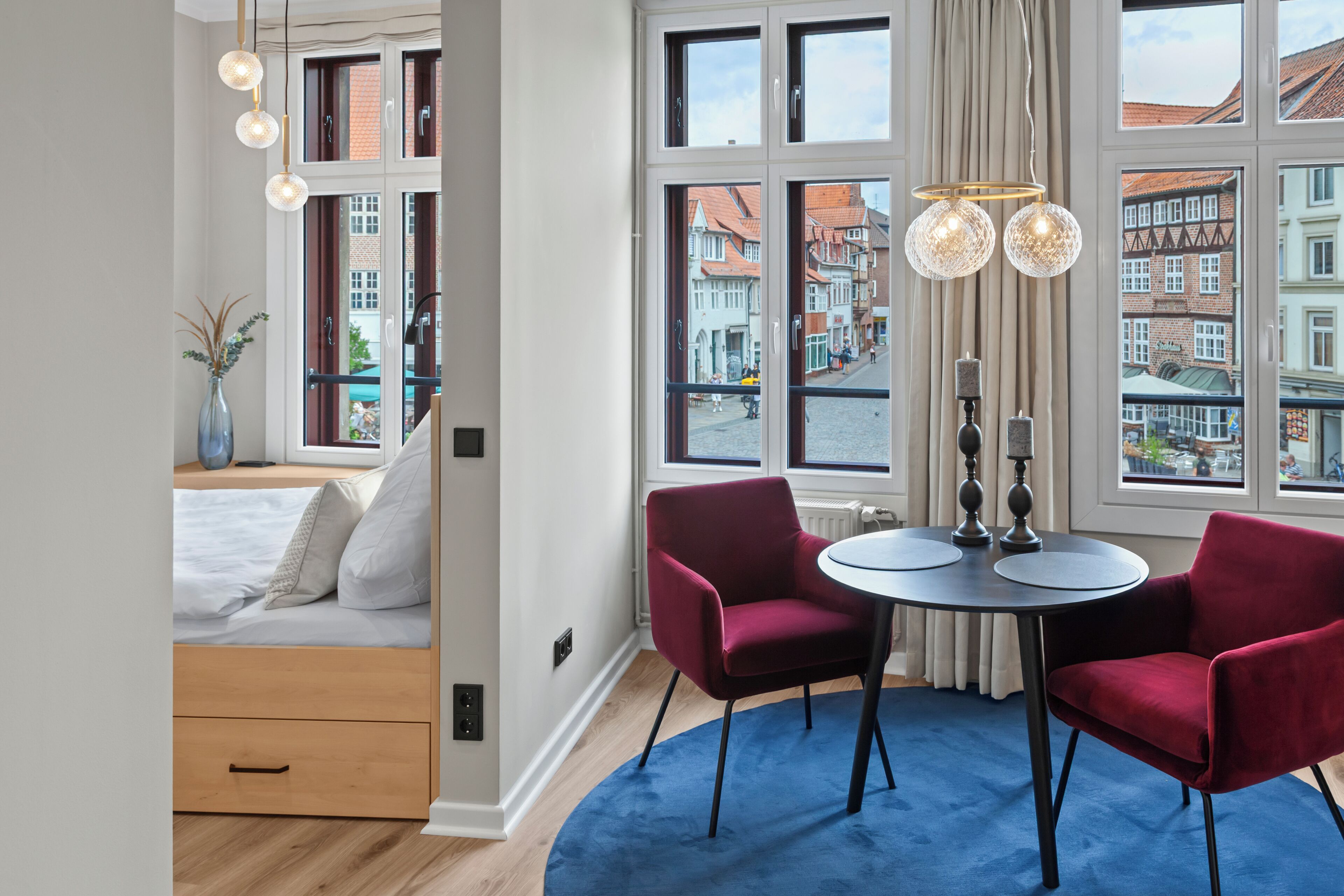A modern hotel room with a bed, a small desk, two red chairs, a round table, and large windows offering a city view.