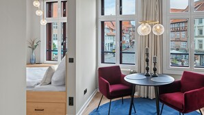 A modern hotel room with a bed, a small desk, two red chairs, a round table, and large windows offering a city view.