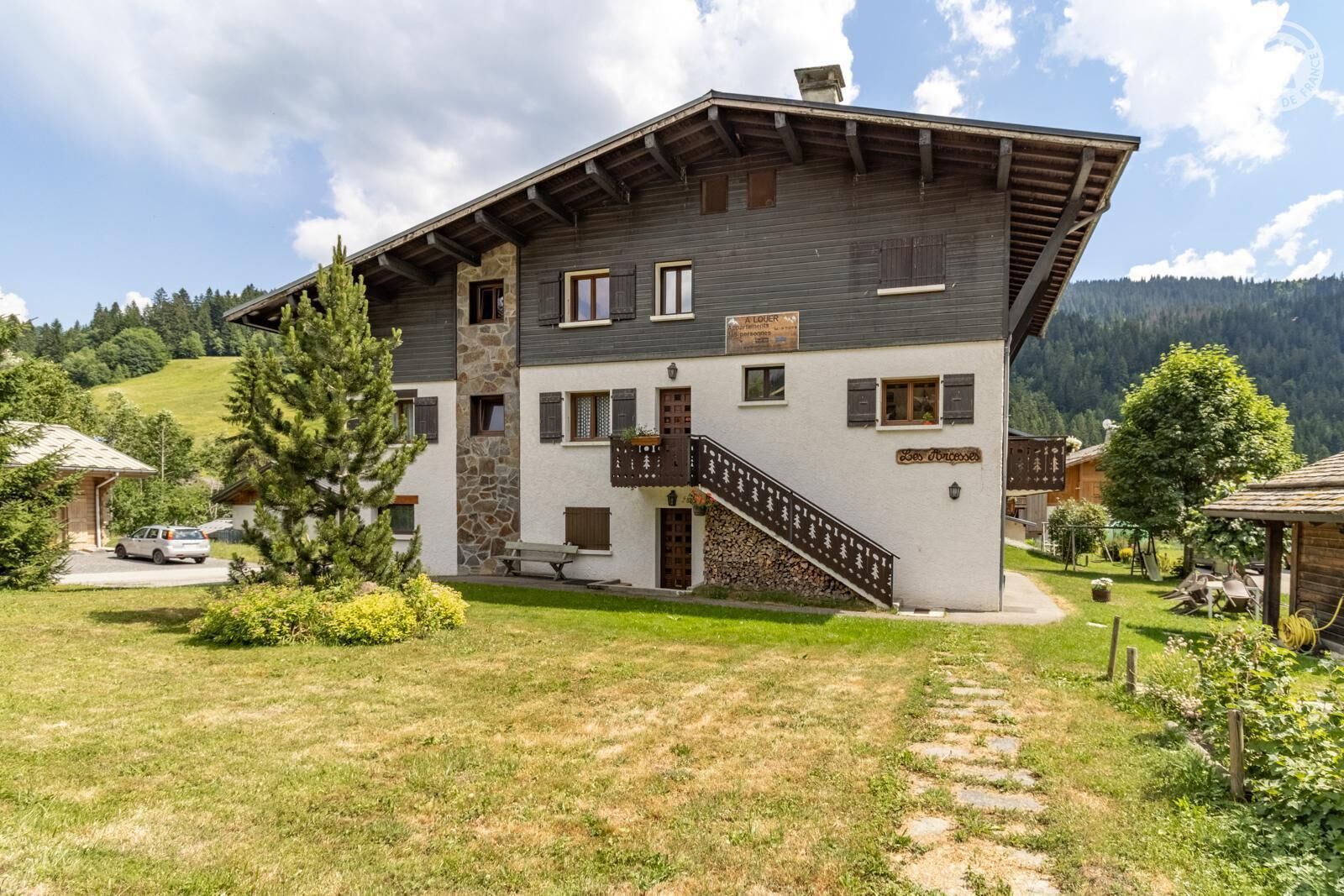 A two-story chalet with a stone facade, a wooden staircase, and a sign reading "Les Jardins".