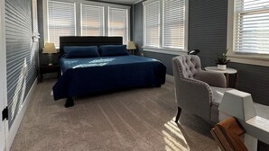 A bedroom with a large bed, a chair, a small table, and a window with blinds.