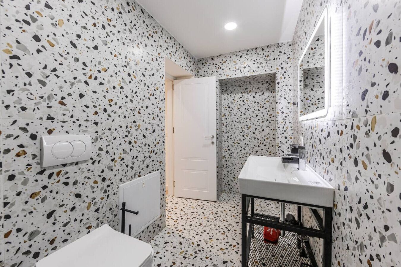 A bathroom with terrazzo flooring, a white sink, a mirror, and a door leading to another room.