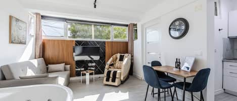 A modern living room with a sofa, a massage chair, a dining area with chairs, a TV, and a clock on the wall.