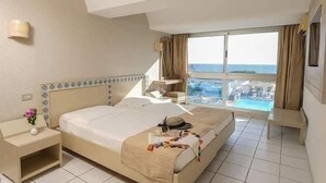 A hotel room with a bed, a bedside table, a nightstand, a window with a view of a pool and buildings, and a ceiling with a skylight. - Hôtel Les Pyramides Club  (Nabeul)