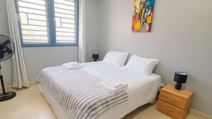 Una cama bien tendida con ropa blanca, una mesita de noche con lámpara y una ventana con cortinas.