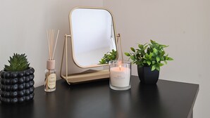 Une coiffeuse noire avec un miroir, une bougie blanche, une petite plante en pot, un diffuseur à bâtonnets et un cœur décoratif accroché au mur.