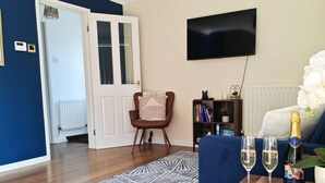 Un salon avec un plancher en bois, un tapis à motifs bleus, un téléviseur, une bibliothèque et une table sur laquelle se trouvent deux verres et une bouteille de champagne.
