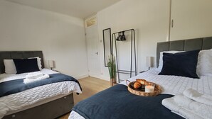 Une chambre d’hôtel avec deux lits, chacun recouvert d’une couvre-lit bleu foncé et doté de draps blancs, une table de chevet avec une lampe et une étagère murale ornée d’objets décoratifs.