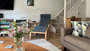 Un salon avec un fauteuil bleu, un canapé gris, une table basse en bois ornée d’un vase de fleurs, un téléviseur à écran plat et une bibliothèque.