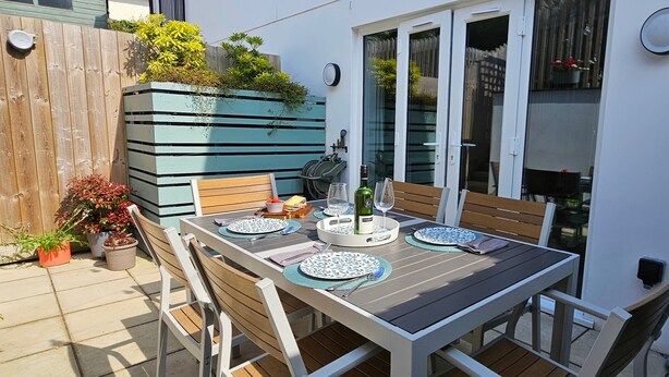 Une terrasse avec une table en bois, dressée pour quatre personnes, entourée de chaises, et un bac à fleurs bleu contenant des plantes.