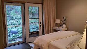 A bedroom with a bed, a nightstand, a lamp, a view through a glass door, and a small table with books.