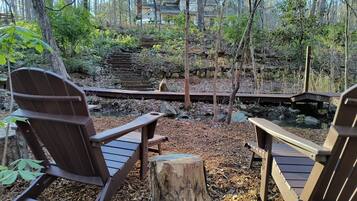 Terraza de madera con sillas Adirondack con vista a un área natural que incluye una escalera de piedra y un pequeño arroyo.