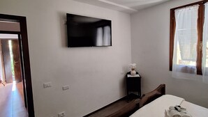 Una habitación de hotel con una cama, un televisor montado en la pared, una mesita de noche con un florero, una ventana con cortinas y un ventilador de techo.