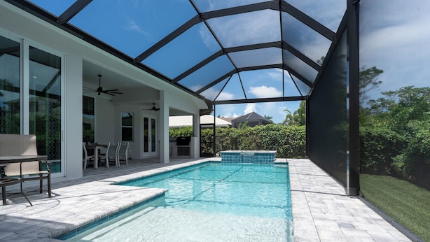 A covered outdoor swimming pool with a patio area, including a table and chairs, and a view of the surrounding greenery. - WALK or BIKE to Beach, HEATED & PRIVATE pool & jacuzzi. NEWLY built & stunning. (Marco Island)