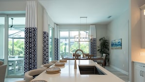 A modern kitchen with a central island, a dining area with chairs, and a large window with curtains. - WALK or BIKE to Beach, HEATED & PRIVATE pool & jacuzzi. NEWLY built & stunning. (Marco Island)