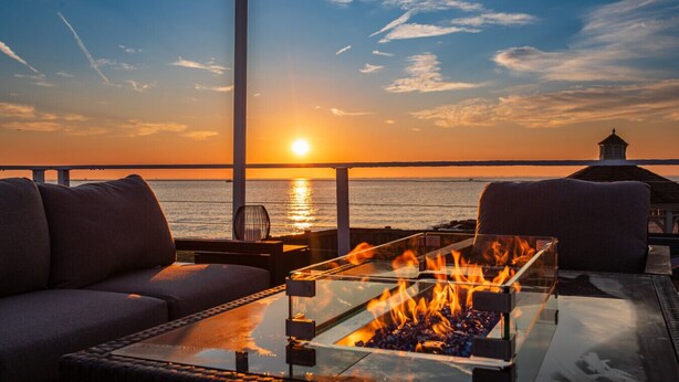 A sunset view from a balcony with a glass-top table and a fire feature, overlooking the sea.