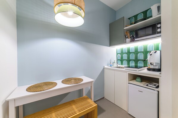 A compact kitchen with a white table, two woven placemats, a wooden bench, a coffee machine, a microwave, and a small refrigerator.