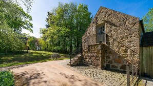 A stone building with a small balcony, a dirt path leading to it, and surrounding greenery. - The Byre, Countryside Home (Tough)