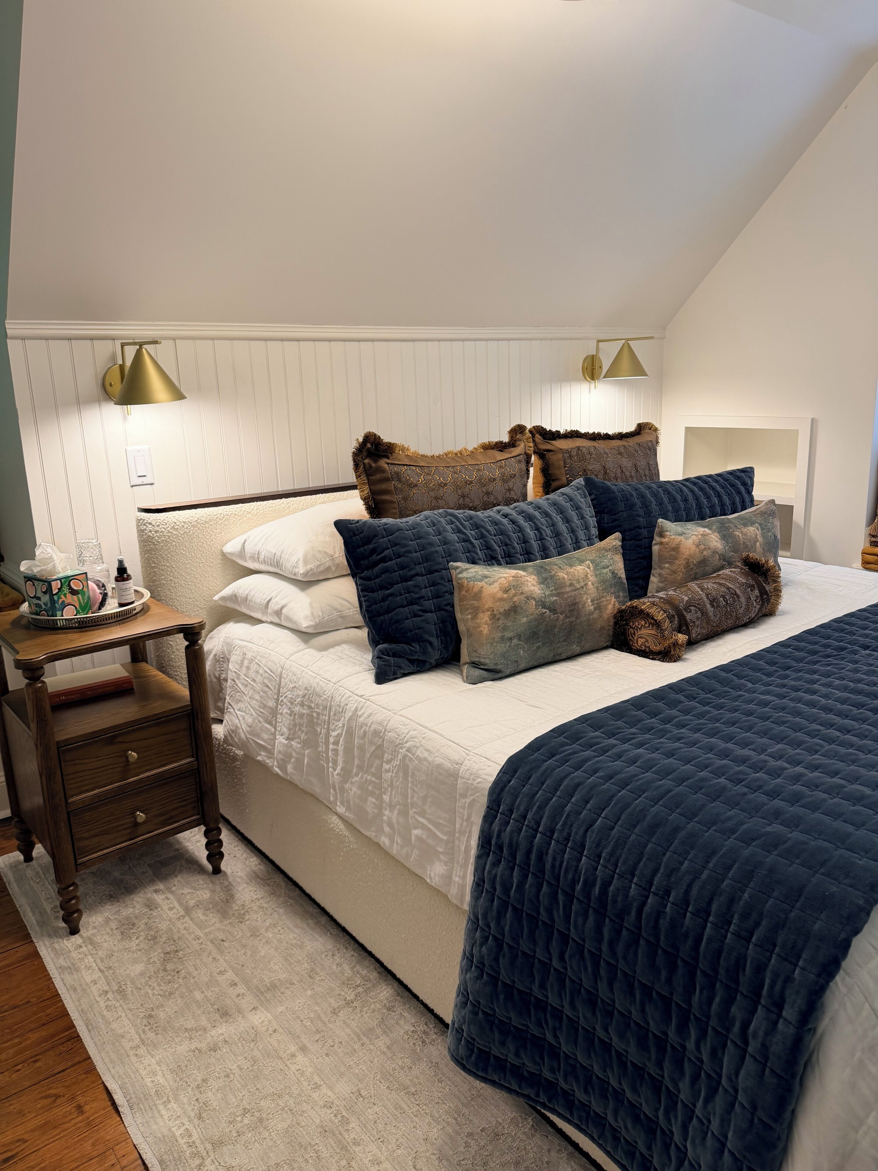Un dormitorio con cama, almohadas, una mesita de noche y una lámpara.