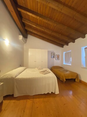 Une pièce avec un plafond en bois, un lit recouvert d’une courtepointe blanche, un petit canapé avec un coussin jaune et une fenêtre munie d’un store.