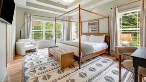 A spacious bedroom with a four-poster bed, a sitting area with a chair, a small table, and a large window offering a view of greenery. - Spacious & Luxe~Hot Tub~Bocce~Fenced Backyard~Mins to Downtown! (Asheville)