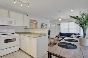 A modern kitchen with white cabinetry, a built-in microwave, and a stove. There is a dining area with a wooden table and blue cushions.