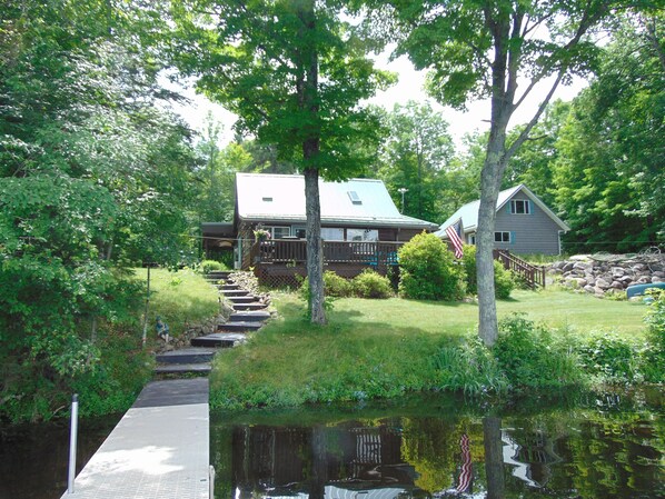 A cabin with a dock extending into a pond, surrounded by lush greenery and trees.
