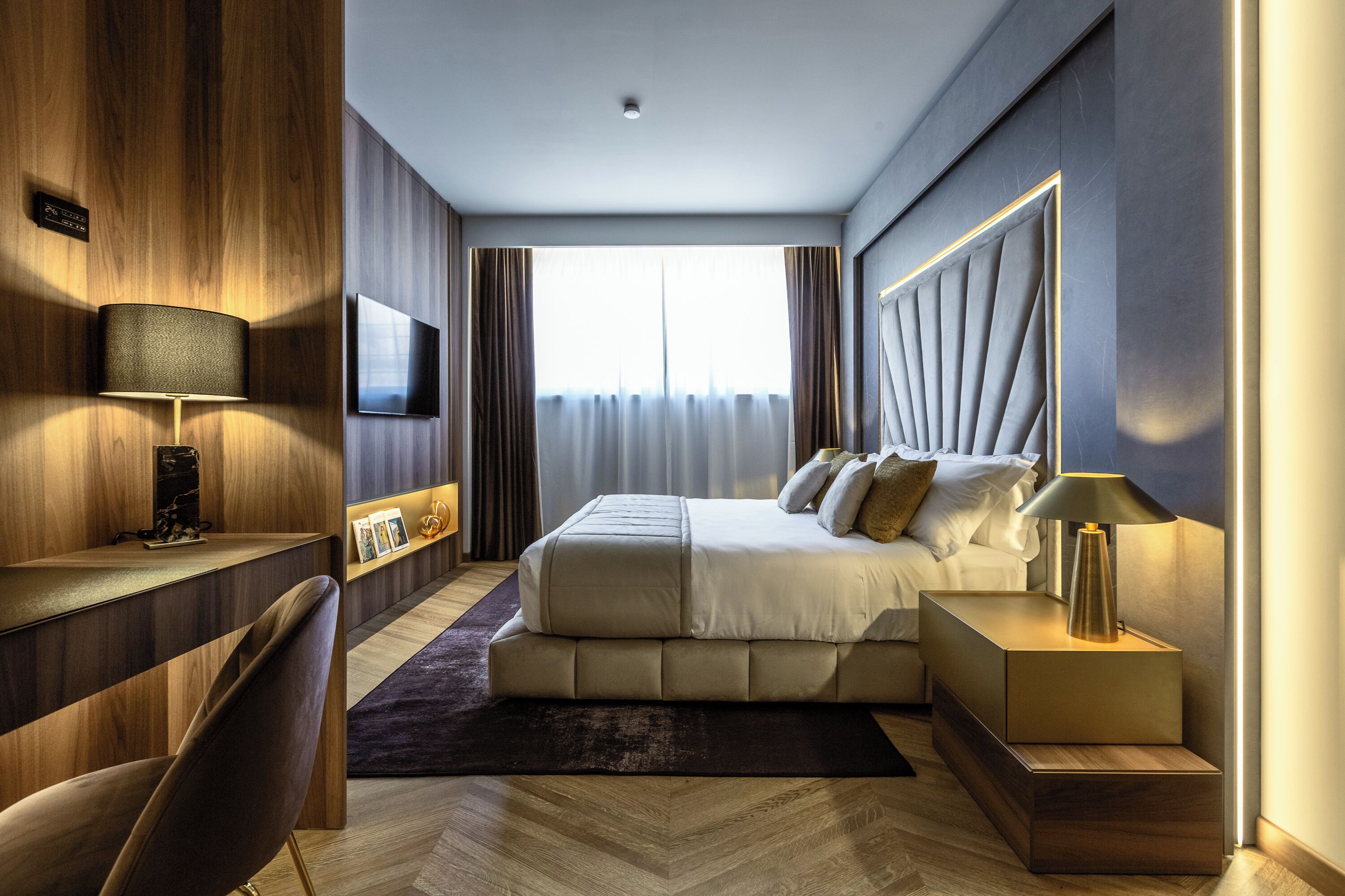 A modern hotel room with a large bed, a desk, a chair, a TV, and a window with curtains.