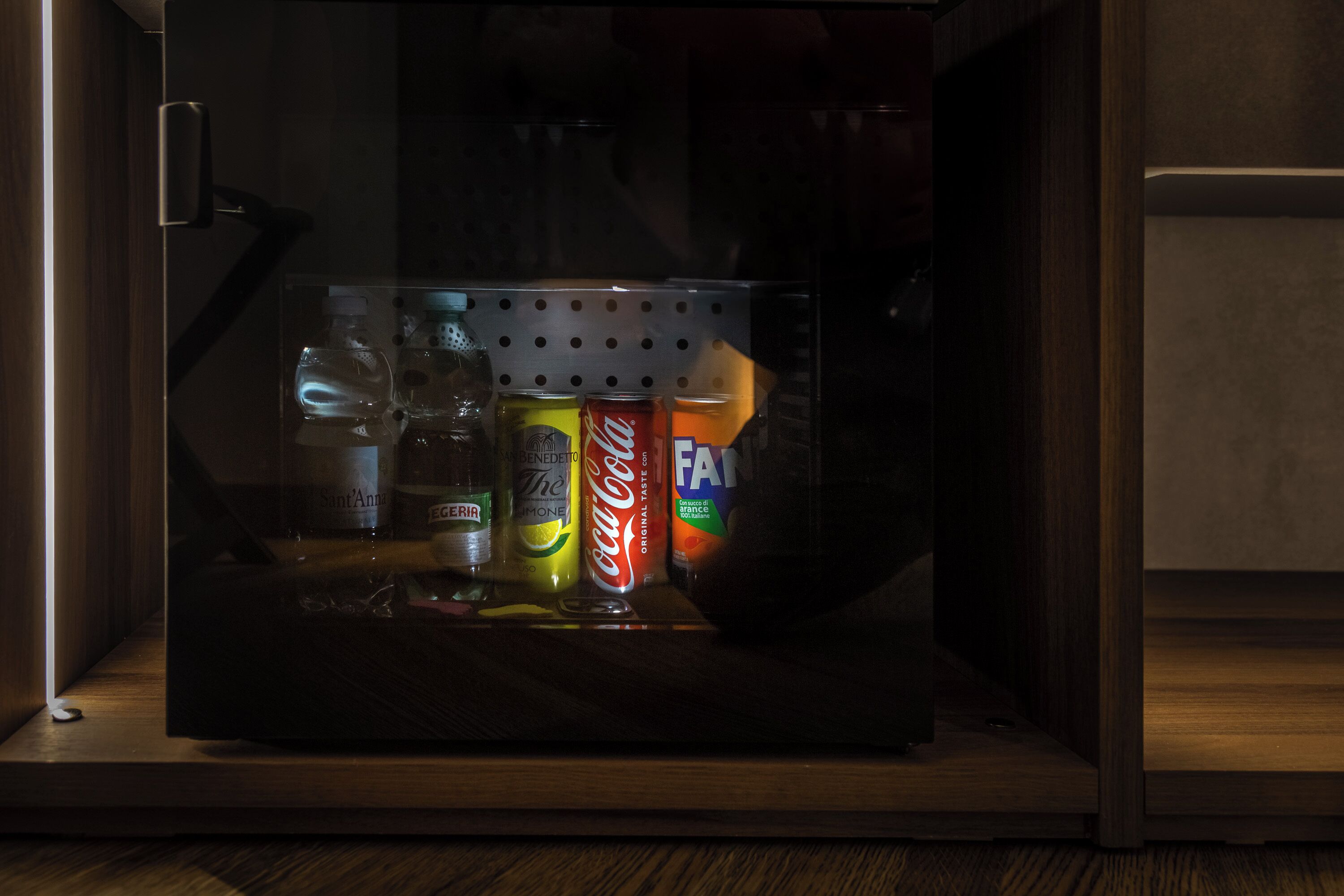 A minibar with various beverages including a bottle of water, a bottle of wine, a bottle of champagne, a can of Coca-Cola, and a can of Fanta.