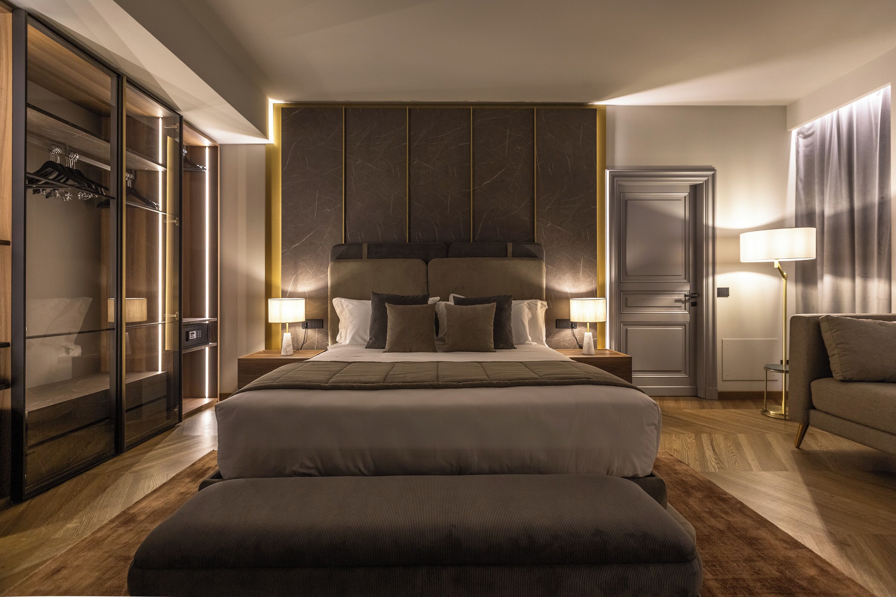 A modern hotel room with a large bed, a wardrobe, a desk, and a seating area.