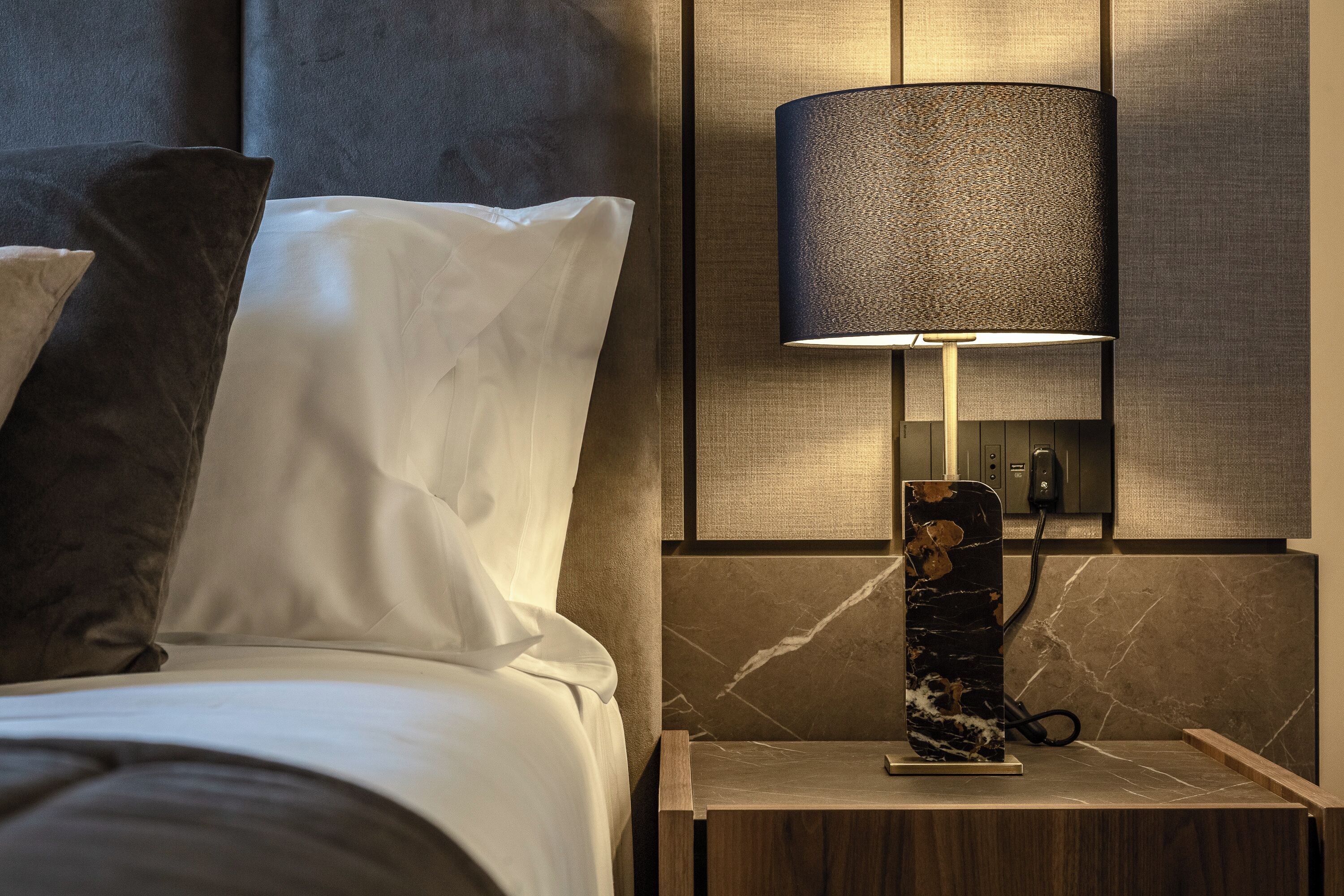 A neatly made bed with white linens and dark pillows, a modern wall-mounted lamp, and a marble-patterned nightstand.