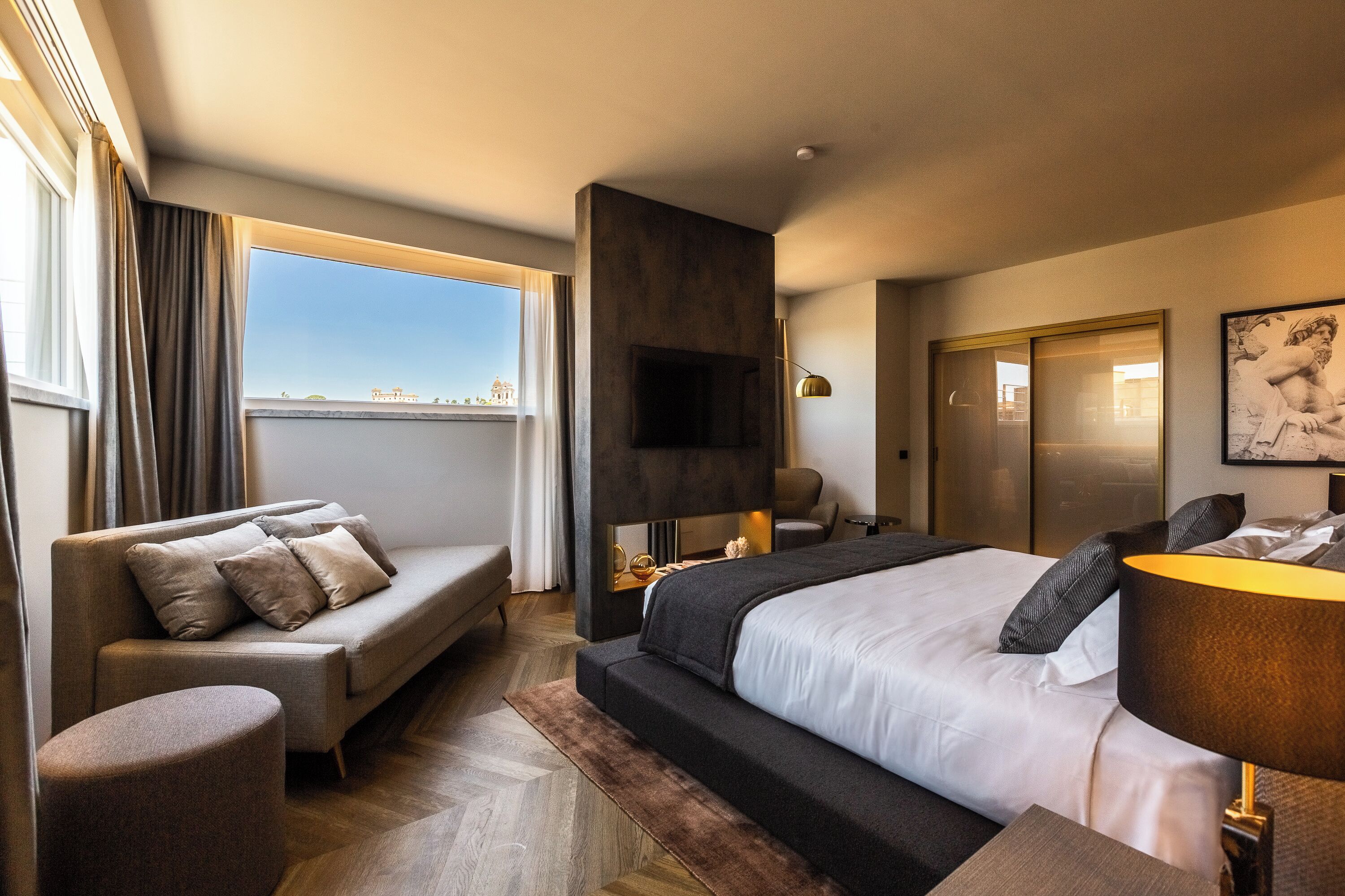A modern hotel room with a large bed, a sofa, a TV, and a city view.