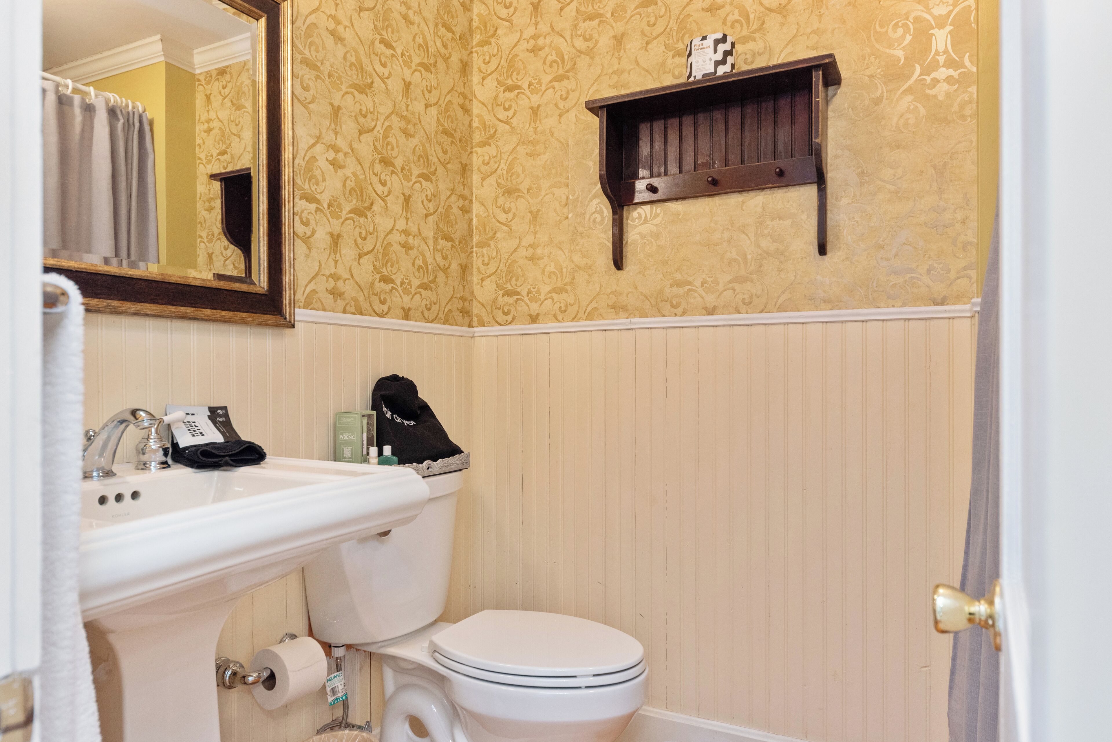 A bathroom with a white sink, toilet, and mirror. There is a towel rack and a shelf above the toilet.