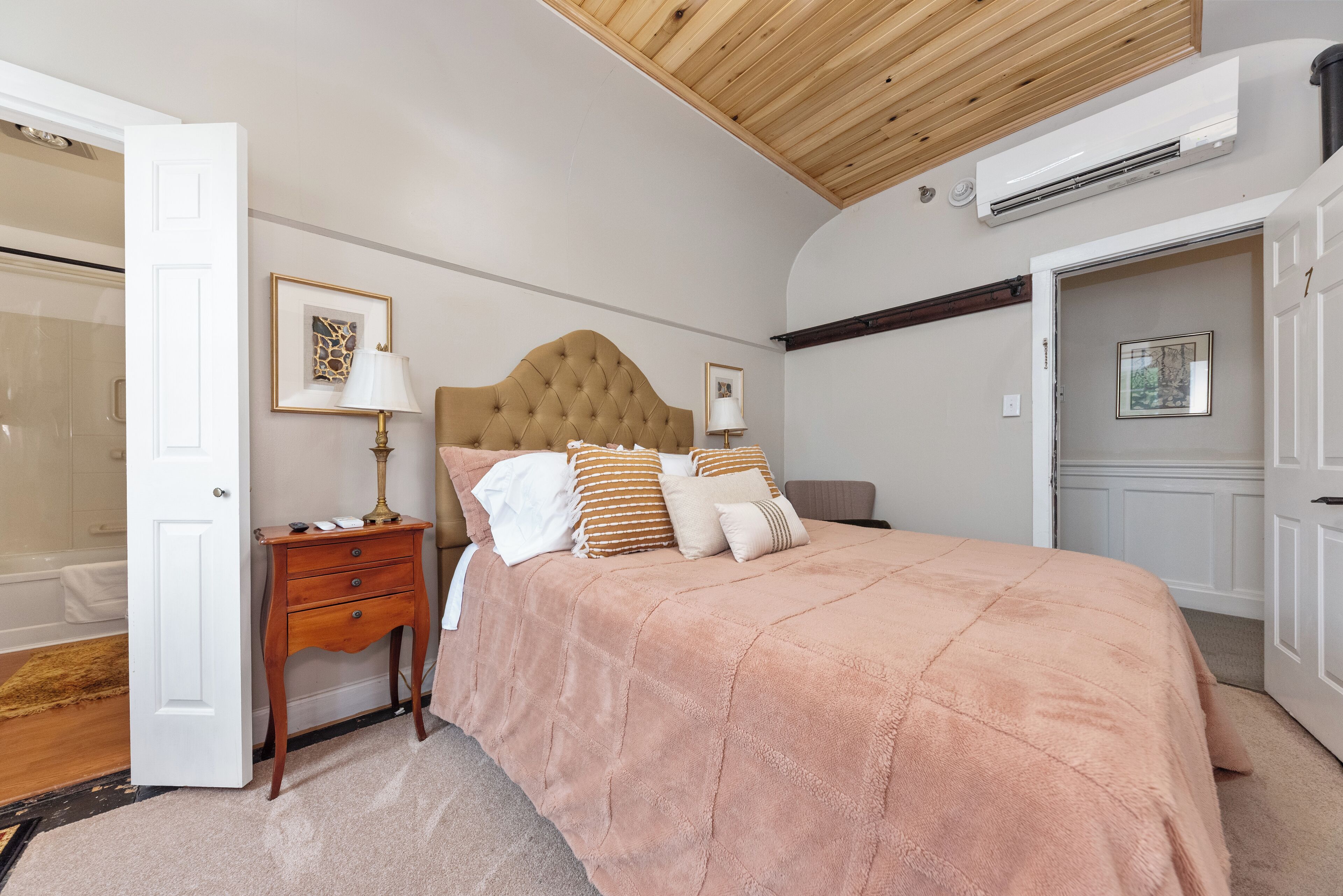 A bedroom with a tufted headboard, a large bed with a peach-colored blanket, a bedside table with a lamp, and a door leading to a bathroom.
