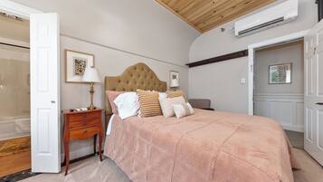 A bedroom with a tufted headboard, a large bed with a peach-colored blanket, a bedside table with a lamp, and a door leading to a bathroom.