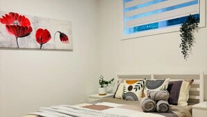 Ein Schlafzimmer mit Bett, Nachttisch, Fenster mit Jalousien, einem Bild und einer hängenden Pflanze.