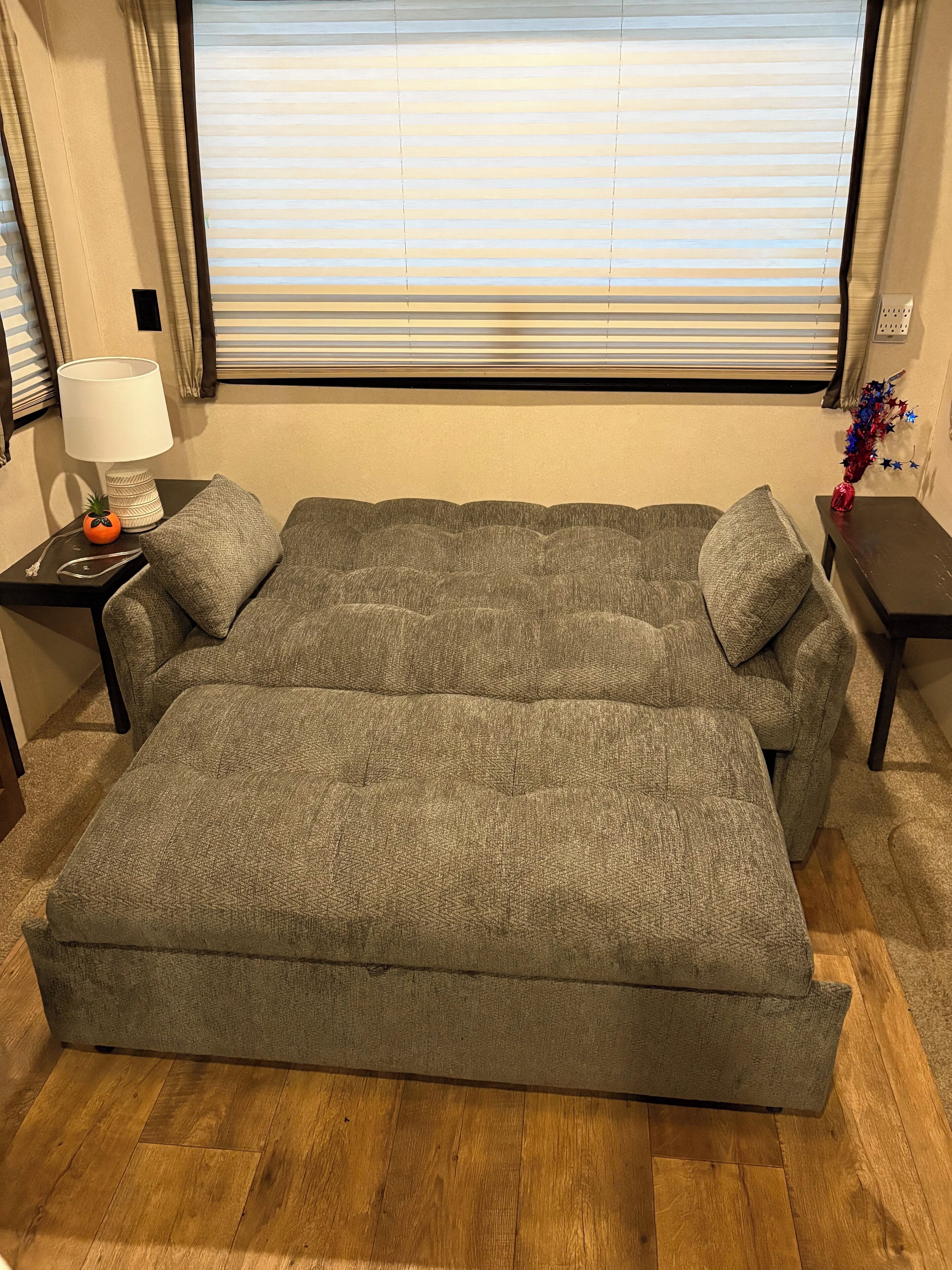 Una habitación con un sofá grande de color gris, una mesita con una lámpara y una ventana con persianas.