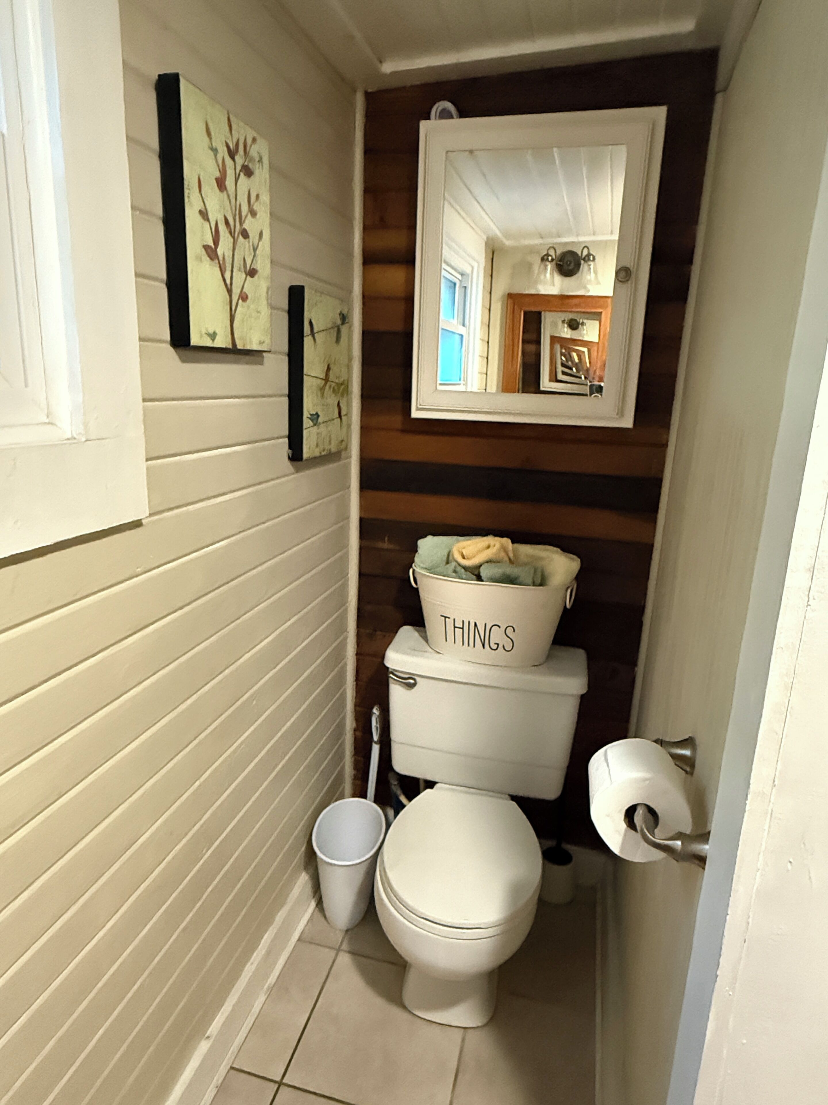Ein kleines Badezimmer mit Toilette, Waschbecken mit einem 'THINGS'-Eimer, Spiegel und einem gerahmten Bild an der Wand.