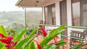 A balcony with a view of a lush landscape, a person sitting on a chair, and a table with a potted plant.