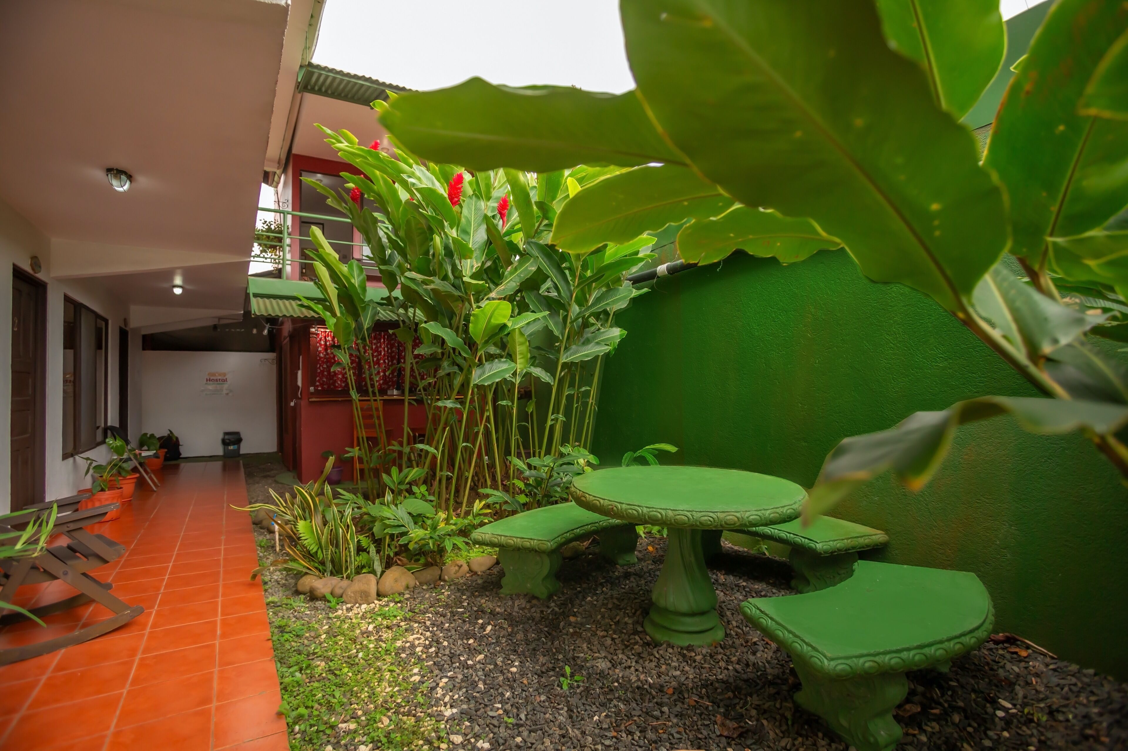 Un patio con una mesa verde y bancos, rodeado de plantas y un muro rojo.