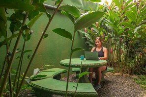 A person sitting at a green table with a laptop, surrounded by lush greenery and plants. - Hostal Nuevo Arenal (Nuevo Arenal)