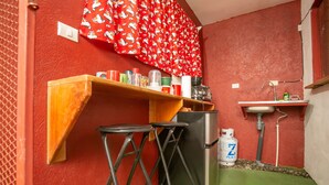 A compact kitchen with a red wall, a wooden shelf with various containers, a refrigerator, a gas cylinder, and a small sink.