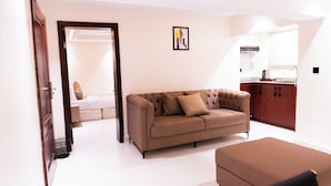 Un salon moderne avec un canapé marron, un pouf blanc et un coin cuisine visible à travers une porte ouverte.