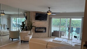 A spacious living room with a large flat-screen TV mounted on a brick wall, a ceiling fan, and a sliding glass door leading to a balcony with outdoor seating.