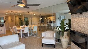 A modern living room with a glass partition, white furniture, a ceiling fan, and a large plant.
