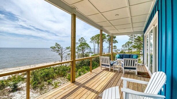 A wooden deck with white Adirondack chairs, a small table, and a view of the ocean.