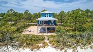 A two-story beachfront house with a balcony, surrounded by dense pine trees and sandy terrain.