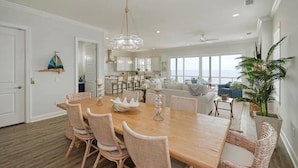 A spacious dining area with a wooden table and wicker chairs, a chandelier, and a view of the ocean through large windows.