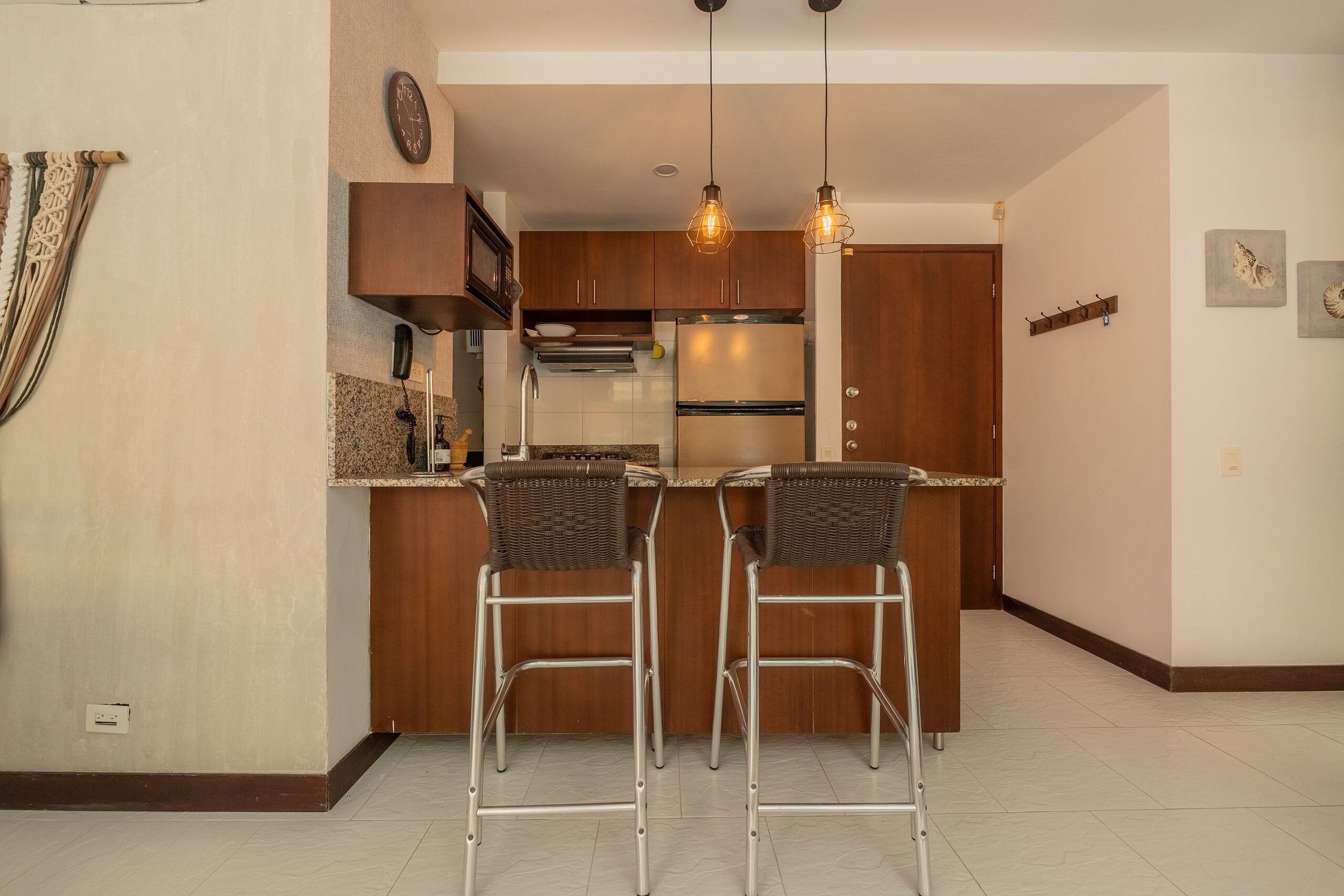 A kitchen with two bar stools, wooden cabinets, a stainless steel refrigerator, and pendant lights.
