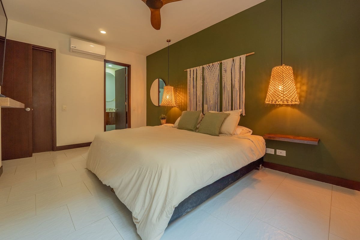 Quarto com cama, ar condicionado na parede, ventoinha no teto e luminária de cabeceira.